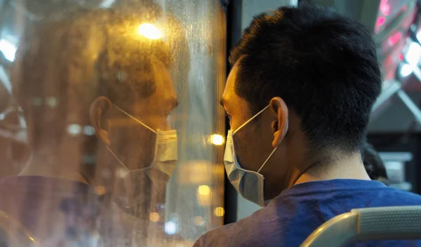 Taichung, Tayvan - 17 Nisan 2020: Bir adam gece boyunca bir otobüsün içinde maske takıyor ve penceresindeki yansıması endişe ve endişe duygularını gösteriyor