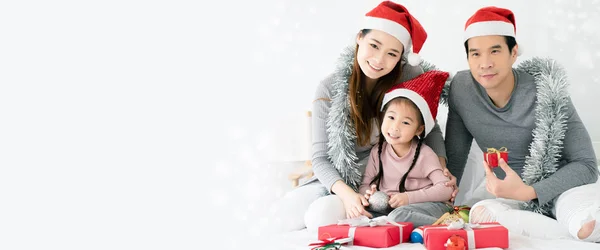 Asyalı ailenin gri, pembe günlük kıyafetleri ve Noel Baba şapkaları olan bir annesi ve kızı var. Kırmızı bir hediye kutusu ve küçük bir topla beyaz bir yatakta oturmak. Noel 'i ve Mutlu Yıllar' ı kutlayın. Boşluğu kopyala