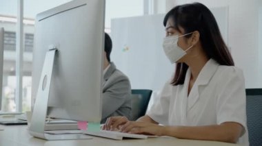 Asyalı işadamları ve kadınlar modern ofiste oturmak ve çalışmak için koruyucu maskeler takarlar. Pazarlama bilgileri üzerinde çalışmak için bilgisayarları kullanın. Koronavirüsü yaymayı bırak. Ya da iş arkadaşları için grip.