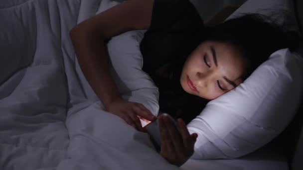 アジアの若い女性がベッドの上に寝そべって夜の時間帯にスマートフォンを再生します 携帯電話で社会を読む 低光で携帯電話を使用すると 目に健康への影響があります 不眠症の概念 — ストック動画