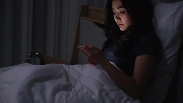アジアの若い女性がベッドの上に寝そべって夜の時間帯にスマートフォンを再生します 携帯電話で社会を読む 低光で携帯電話を使用すると 目に健康への影響があります 不眠症の概念 — ストック動画