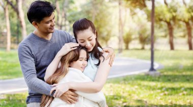 Asyalı ailenin anne ve babası parkta gülümseyerek mutlu mesut kızlarına sarılıyorlar. Uzun yaz tatillerinde büyük ağaçlarla. Uluslararası Aile Günü. Sağlık sigortası kavramı