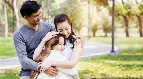 Asyalı ailenin anne ve babası parkta gülümseyerek mutlu mesut kızlarına sarılıyorlar. Uzun yaz tatillerinde büyük ağaçlarla. Uluslararası Aile Günü. Sağlık sigortası kavramı