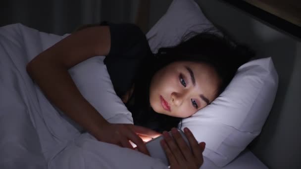 アジアの若い女性がベッドの上に寝そべって夜の時間帯にスマートフォンを再生します 携帯電話でCovid 19をお読みください 低光の中でP電話を使用すると 目に健康影響を与えます 不眠症の概念 — ストック動画