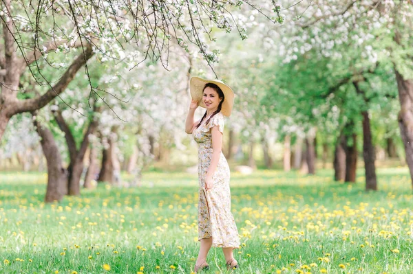 Jovem com chapéu de palha caminha no jardim florido na primavera. Menina debaixo da árvore de flores Sakura. Conceito de moda verão — Fotografia de Stock