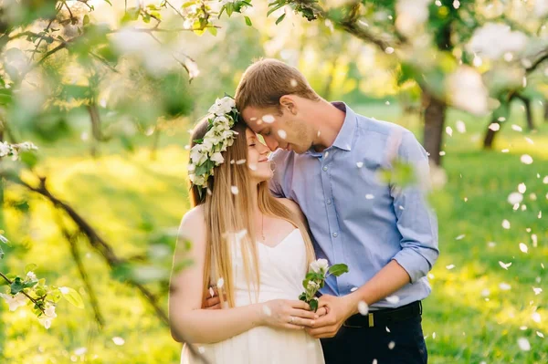 Una donna incinta con un vestito bianco e una corona floreale in testa che sta con il marito in giardino. Si tengono per mano con petali pancia incinta stanno cadendo sotto il sole splendono . — Foto Stock