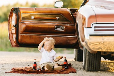 Buick Riviera 'nın retro tarzı. Benzersiz bir araba. Tatlı sarışın çocuk eski arabanın yanında oturuyor, pizza yiyor ve kola içiyor, alnını sıkıca tutuyor.