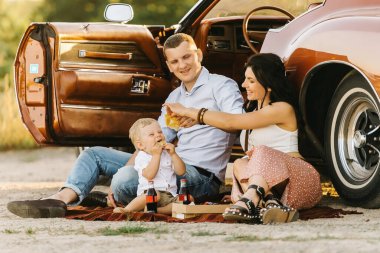 Buick Riviera 'nın retro tarzı. Benzersiz bir araba. Güzel ailesiyle gün batımı pikniğinde retro arabanın yanında oturan sevimli sarışın çocuk pizza yiyip kola içiyor..