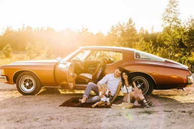 Buick Riviera 'nın retro tarzı. Benzersiz bir araba. Güzel ailesiyle gün batımı pikniğinde retro arabanın yanında oturan sevimli sarışın çocuk pizza yiyip kola içiyor.