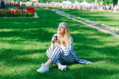 Şanslı bir kadın yeşil çimenlerde pijamaları ve uyku maskesiyle oturur ve güneşin tadını çıkarır ve kahve içer..