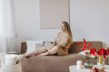 İçi bej kaplamalı ve şık bej takım elbiseli güzel bir kadın kitap okuyor, kırmızı çiçekler. Ev ve konfor kavramları