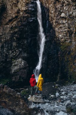 Gürültü efekti, seçici odak: parlak renkli yağmurluk giymiş minyatür turist şelaleli bir uçurumun geniş ölçekli arka planında çelişkili bir şekilde görülüyor