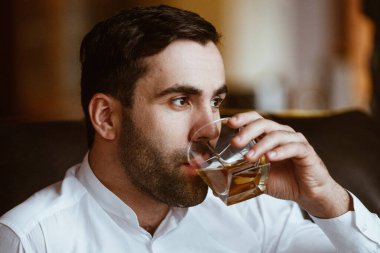 Şık, karizmatik bir adam elinde bir bardak alkolle büyük kır evinde bir sandalyede oturuyor.