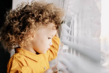 01.06.2019 Vinnitsa, Ukrayna: Kıvırcık bebek parlak güneş ışığıyla pencereden dışarı bakar.