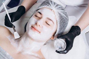 Yüzünde spa ameliyatı olan genç bir kadın. Spa masasına uzanmış bir kadın ve kozmetik maske takan bir kozmetik uzmanı. Metnin için boşluk kopyala