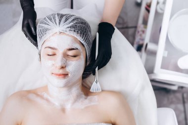 Yüzünde spa ameliyatı olan genç bir kadın. Spa masasına uzanmış bir kadın ve kozmetik maske takan bir kozmetik uzmanı. Metnin için boşluk kopyala