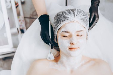Yüzünde spa ameliyatı olan genç bir kadın. Spa masasına uzanmış bir kadın ve kozmetik maske takan bir kozmetik uzmanı. Metnin için boşluk kopyala