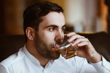 Şık, karizmatik bir adam elinde bir bardak alkolle büyük kır evinde bir sandalyede oturuyor.