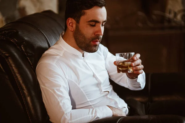stylish charismatic man with a glass of strong alcohol in his hands sits on a chair in his large country house