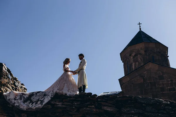 stylish Georgian groom dressed in a national costume leads his bride holding her hand on a rocky cliff against the backdrop of the sky and the church where the wedding was held