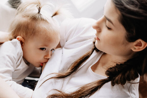 молодая красивая женщина кормит грудью свою маленькую дочь, сидя на подоконнике своей спальни
