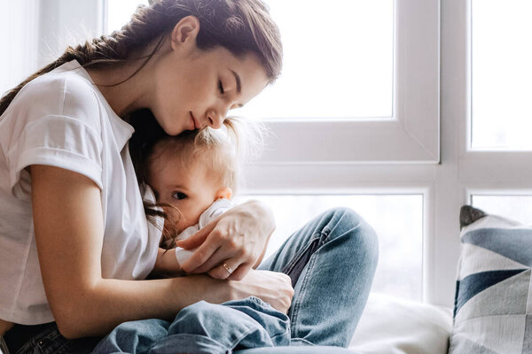 молодая красивая женщина кормит грудью свою маленькую дочь, сидя на подоконнике своей спальни
