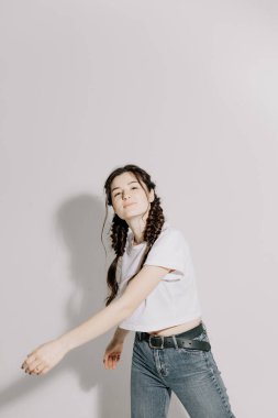 Beyaz tişört ve kot pantolon giymiş atölye ışığı önünde beyaz arka planda fotoğraf için poz veren at kuyruklu güzel bir kadın.