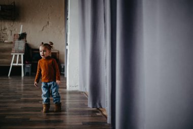 2 yaşındaki küçük çekici kız, büyük bir stüdyoda, penceresinin yanında perdeleri olan bir fotoğraf için poz veriyor.