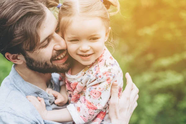 公園の散歩中の緑の木々を背景に小さな娘を腕に持つスタイリッシュなひげそりの父親抱擁し 赤ちゃんにキス — ストック写真