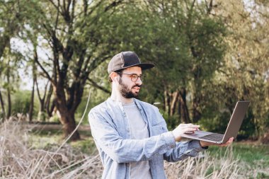 Gözlüklü, koyu renk saçlı çekici genç adam güneşli bir günde dışarıda dizüstü bilgisayarla çalışıyor. Sakallı hippi adam günlük kıyafetler giyiyor ve karantina altında çalışıyor.