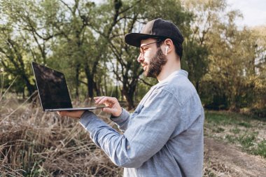 Genç hippi dışarıda dizüstü bilgisayar kullanıyor. Güneşli yaz gününde parkta bilgisayarla çalışan yakışıklı bir adam. Açık hava doğa yolculuğu ve rahatlama. Serbest çalışma kavramı.