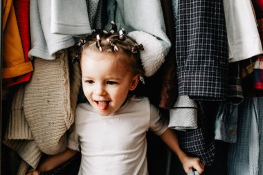 Askıda asılı kıyafetlerin arkasına saklanan bir çocuğun portresi. Karantina oyunları, ebeveynler çocuklarıyla saklambaç oynuyor. Ev yapımı saklambaç oyunu.