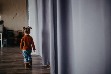 2 yaşındaki küçük çekici kız, büyük bir stüdyoda, penceresinin yanında perdeleri olan bir fotoğraf için poz veriyor.