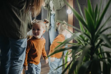 2 yaşındaki küçük çekici kız aynanın yanında makyaj yapan annesinin yanında oynuyor, bebek de annesinin aynanın önünde poz vermesini izliyor.