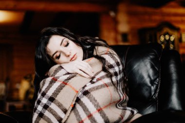 Sıcak bir battaniyeye sarılı genç ve çekici bir kadın büyük bir sandalyeye oturur, sarılır ve ısınır.