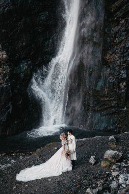Gürültü efekti, seçici odak, manken çekimi: Şelale arka planında çok güzel uzun bir elbise ve damat giymiş çekici bir gelin.