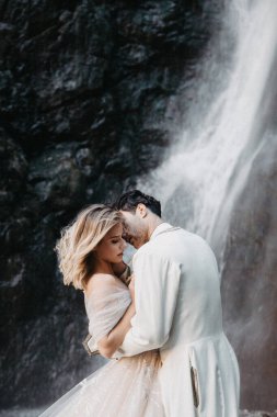 Gürültü etkisi, seçici odak: samimi duygular, tutku ve gelinlerin aşkı dağlarda bir düğün fotoğrafı çekiminde kayaların ve şelalenin arka planına karşı