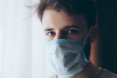 Tıbbi maske takan genç bir adamın portresi. Evde ya da pencerenin karşısındaki hastane koğuşunda karantinaya alınmış.