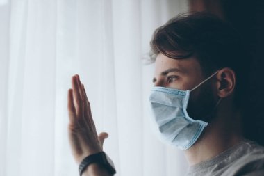 Salgın. Corona virüsünün hasta adamı pencereden dışarı bakıyor ve maske takıyor ve evdeki hastalıktan korunuyor..
