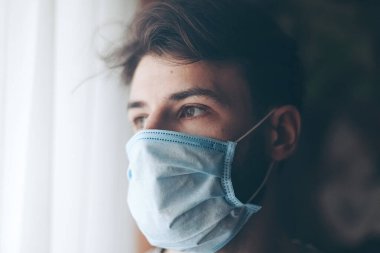 Coronavirus karantinasında koruyucu maske takmış bir adam camdan dışarı bakıyor.