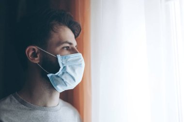 Tıbbi maske takan genç bir adamın portresi. Evde ya da pencerenin karşısındaki hastane koğuşunda karantinaya alınmış.