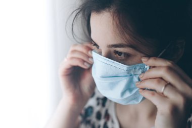 Koruyucu tıbbi maske takan bir kızın portresi. Karantina sırasında pencereden dışarı bakan bir kadın.