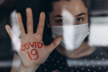 Evdeki karantina altındaki genç kadın avucundaki yazıyla pencereden dışarı bakıyor: COVID 19, koronavirüs enfeksiyonu uyarısı anlamına geliyor.