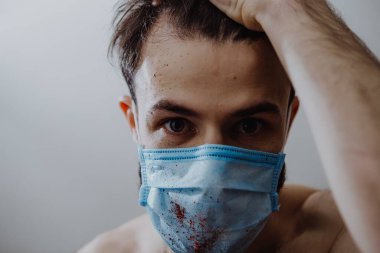Hastalıklı, koruyucu bir maske takan genç bir adam. Kan sıçramış. Bu da hastanın sağlığının bozulduğunu gösteriyor.