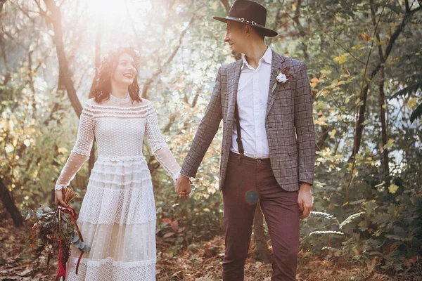 stylish couple of brides: man in tuxedo and hat, woman in light white dress with wreath on head against evening park background, lovers posing at wedding photo shoot. wedding background