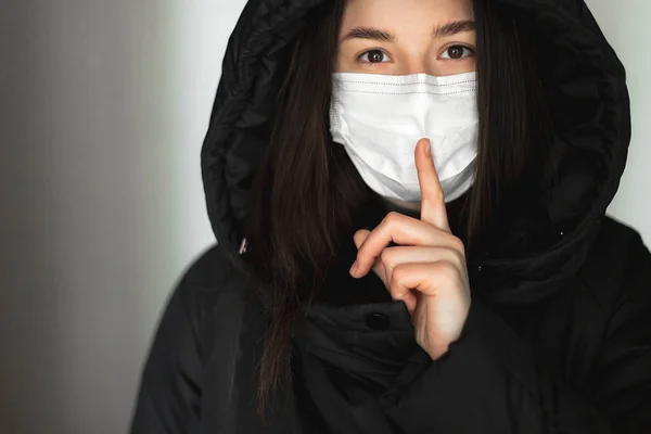 a young woman wearing a medical mask shows a hand sign that means it's a secret and needs to be silent. Medical concept of spread of viral diseases and quarantine