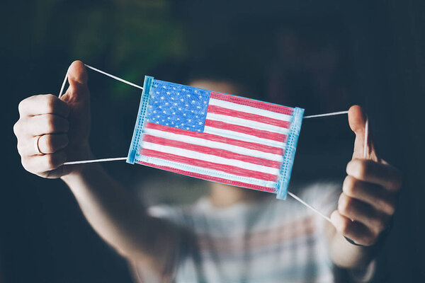 медицинская маска в руках человека, с нарисованным флагом Соединенных Штатов Америки. Копирование пространства
