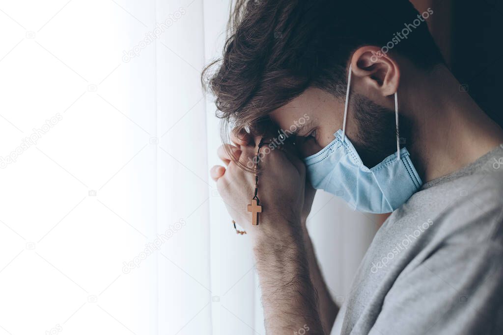 portrait of a young man wearing a medical mask and quarantined at home or in a hospital ward opposite a window with rosary