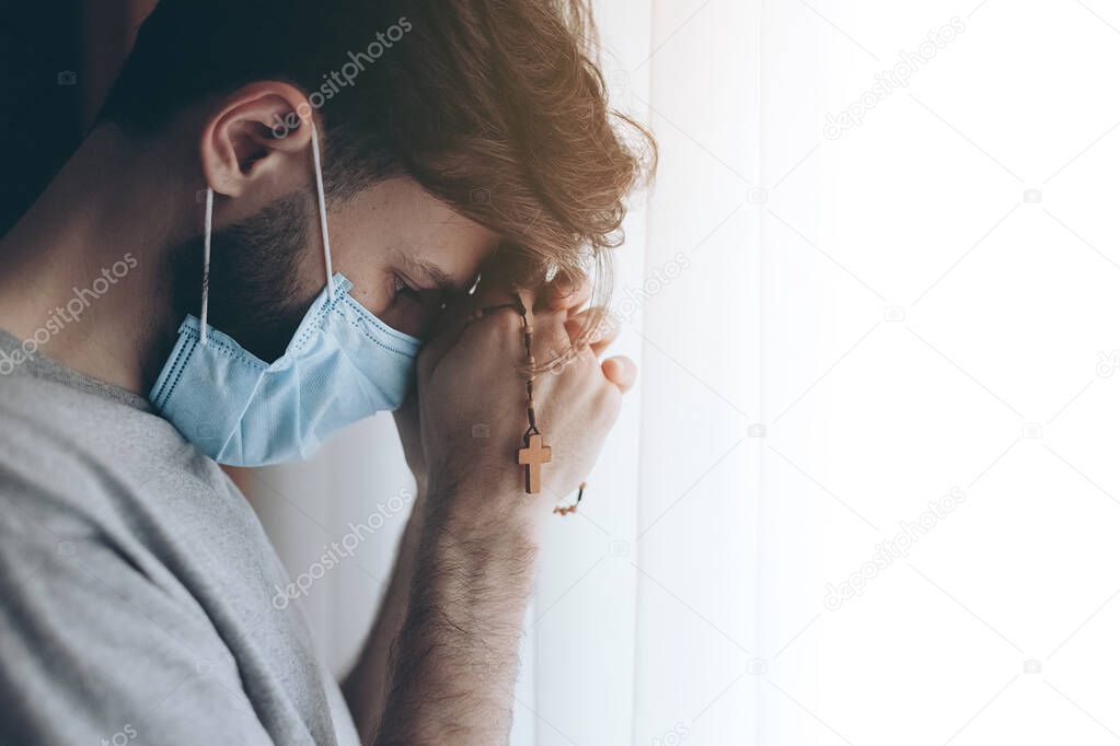 portrait of a young man wearing a medical mask and quarantined at home or in a hospital ward opposite a window with rosary