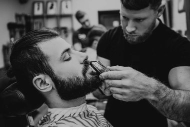 Sakal ve kuaförde kuaförlük yaparken müşterinin portresi. Sakallı berber, bir kesiciyle müşterinin sakalının şeklini oluşturur. Seçici odak, ses efekti